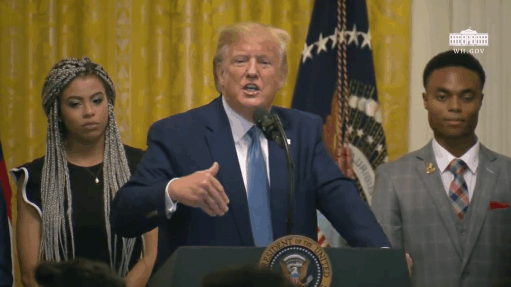 President Donald Trump stands at a podium with the presidential seal, speaking into a microphone. He gestures with his right hand, flanked by two individuals in formal attire. An American flag and a gold curtain are visible in the background, along with the White House insignia.