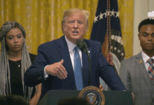 President Donald Trump stands at a podium with the presidential seal, speaking into a microphone. He gestures with his right hand, flanked by two individuals in formal attire. An American flag and a gold curtain are visible in the background, along with the White House insignia.