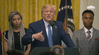 President Donald Trump stands at a podium with the presidential seal, speaking into a microphone. He gestures with his right hand, flanked by two individuals in formal attire. An American flag and a gold curtain are visible in the background, along with the White House insignia.