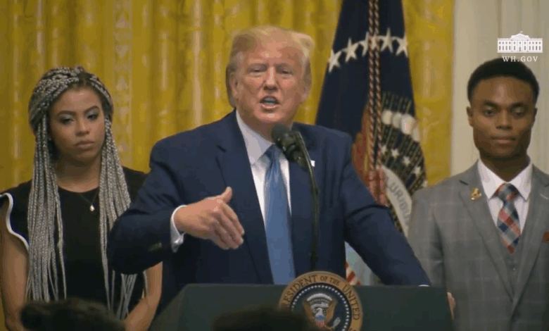 President Donald Trump stands at a podium with the presidential seal, speaking into a microphone. He gestures with his right hand, flanked by two individuals in formal attire. An American flag and a gold curtain are visible in the background, along with the White House insignia.