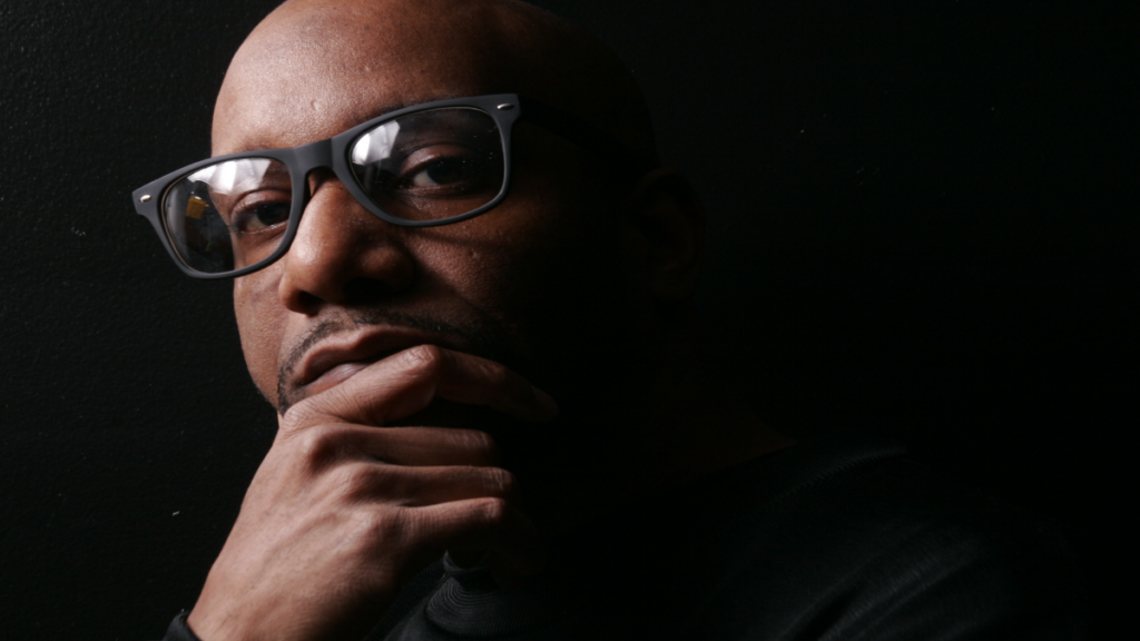 Rahiem Shabazz, Atlanta filmmaker and author of Radical Hope in the 21st Century, in thoughtful portrait wearing dark-framed glasses, hand on chin, black shirt against black background.