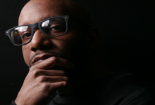 Rahiem Shabazz, Atlanta filmmaker and author of Radical Hope in the 21st Century, in thoughtful portrait wearing dark-framed glasses, hand on chin, black shirt against black background.