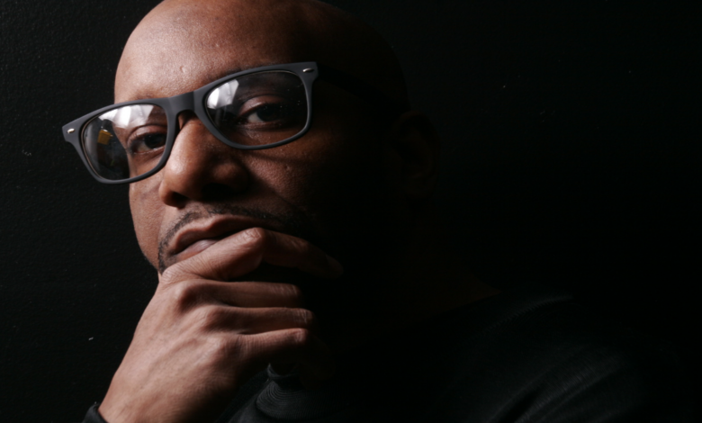 Rahiem Shabazz, Atlanta filmmaker and author of Radical Hope in the 21st Century, in thoughtful portrait wearing dark-framed glasses, hand on chin, black shirt against black background.