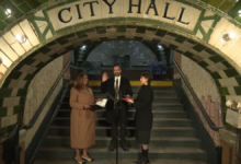Under a tiled archway reading “City Hall” in an old subway station, New York State Attorney General Letitia James stands at left holding papers, Zohran Mamdani stands center with his right hand raised to take the oath of office, and another person stands at right holding a book for him on the steps behind a microphone