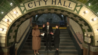 Under a tiled archway reading “City Hall” in an old subway station, New York State Attorney General Letitia James stands at left holding papers, Zohran Mamdani stands center with his right hand raised to take the oath of office, and another person stands at right holding a book for him on the steps behind a microphone