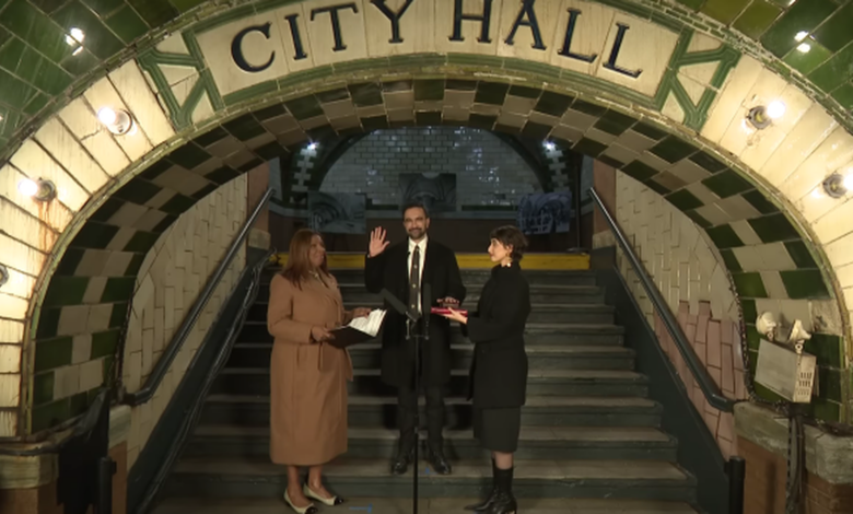 Under a tiled archway reading “City Hall” in an old subway station, New York State Attorney General Letitia James stands at left holding papers, Zohran Mamdani stands center with his right hand raised to take the oath of office, and another person stands at right holding a book for him on the steps behind a microphone
