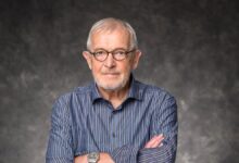 Francis Halzen wearing glasses and a striped shirt, with his arms crossed against a gray background.